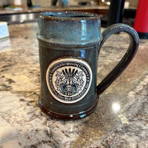 May include: A cylindrical ceramic beer mug with a dark blue and brown reactive glaze finish. The front features a circular cream-colored emblem with the text Swashbuckler Brewing Company, Live Free, Dine Well, Drink Good Ale, and SBC.
