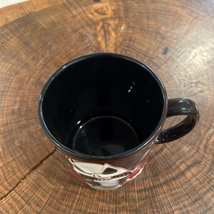 May include: Black ceramic mug with a handle, featuring a decorative design around the rim. The mug is set against a wooden surface. The interior of the mug is black.