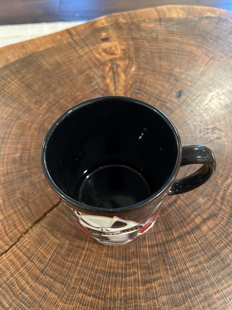 May include: Black ceramic mug with a handle, featuring a decorative design around the rim. The mug is set against a wooden surface. The interior of the mug is black.