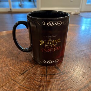 May include: Black ceramic mug featuring the text "Tim Burton's The Nightmare Before Christmas" in gold and red. The mug has a black handle and a decorative white pattern near the rim. The mug is on a wooden surface.
