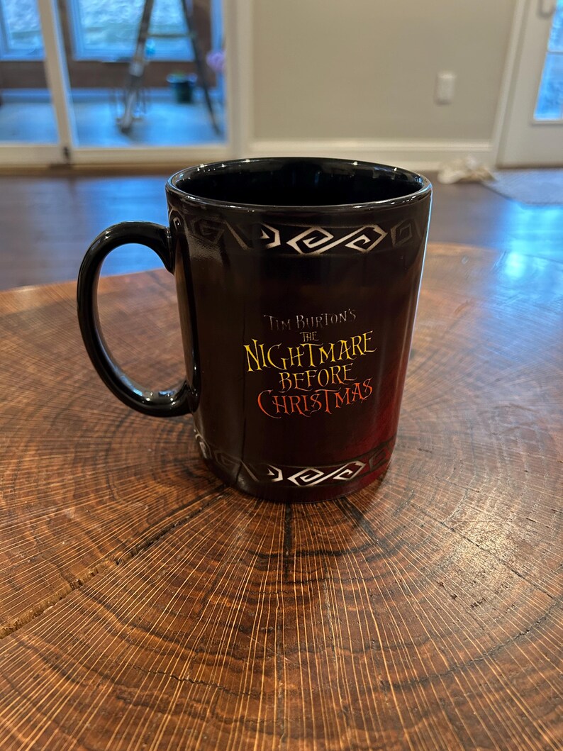 May include: Black ceramic mug featuring the text "Tim Burton's The Nightmare Before Christmas" in gold and red. The mug has a black handle and a decorative white pattern near the rim. The mug is on a wooden surface.