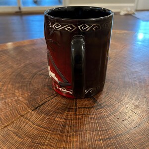 May include: A black ceramic mug with a black handle. The mug features a design with a red background and white accents. The top rim has a silver geometric pattern. The mug is sitting on a wooden surface.