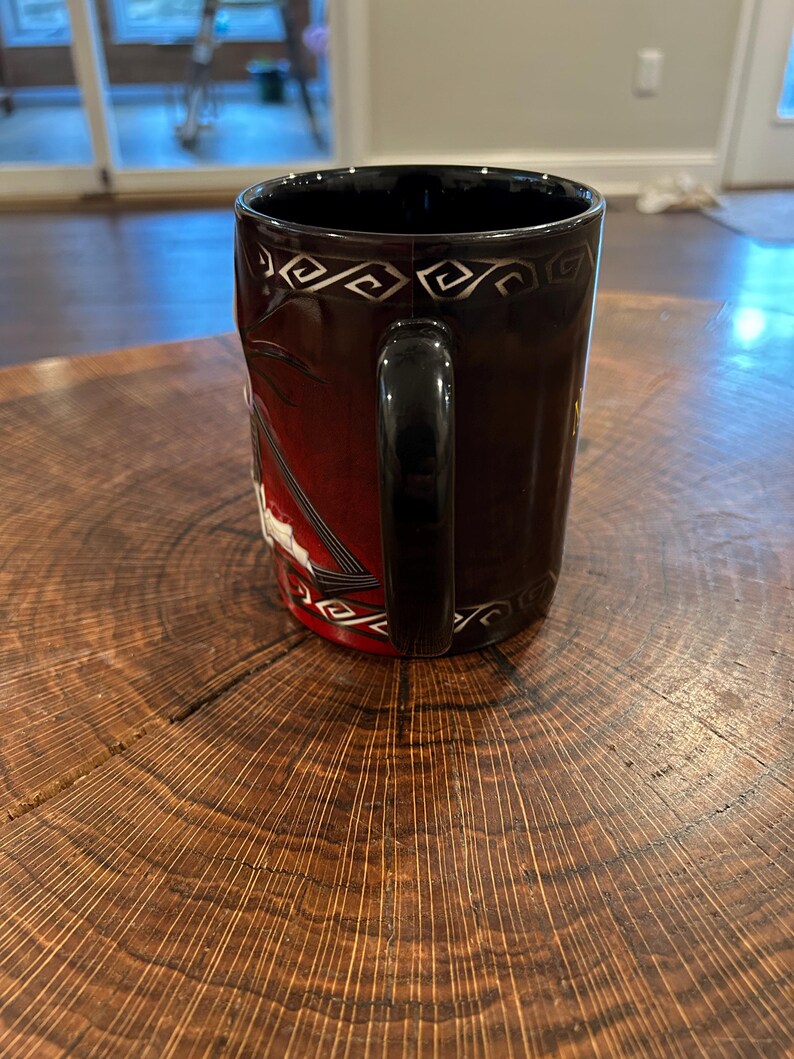 May include: A black ceramic mug with a black handle. The mug features a design with a red background and white accents. The top rim has a silver geometric pattern. The mug is sitting on a wooden surface.