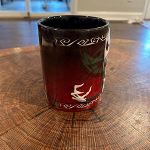 May include: A cylindrical coffee mug with a black interior and a red exterior that fades to black. The mug features a white skeletal hand design and a decorative white border. The mug is sitting on a wooden surface.