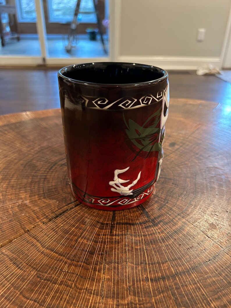 May include: A cylindrical coffee mug with a black interior and a red exterior that fades to black. The mug features a white skeletal hand design and a decorative white border. The mug is sitting on a wooden surface.