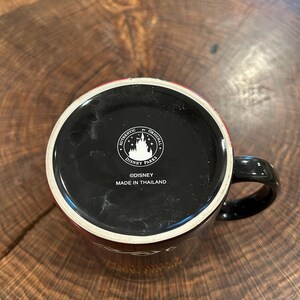 May include: Black ceramic mug with a black handle and a red rim. The bottom of the mug features the text "AUTHENTIC ORIGINAL DISNEY PARKS" and "&copy;DISNEY MADE IN THAILAND". The mug is sitting on a wooden surface.