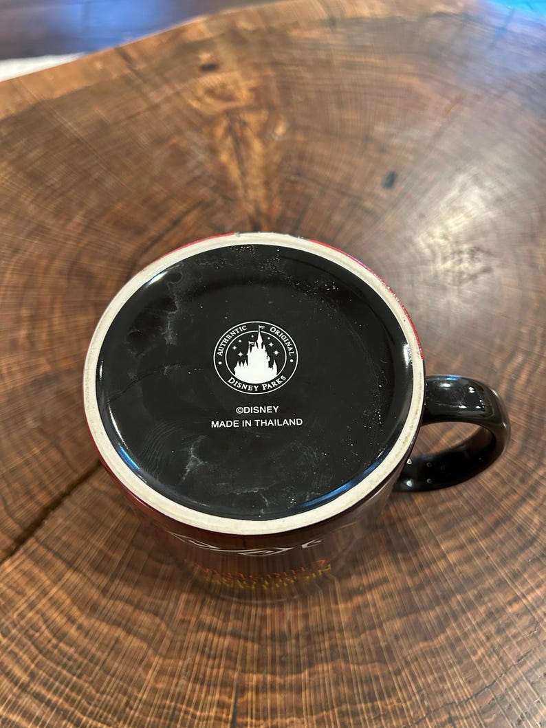 May include: Black ceramic mug with a black handle and a red rim. The bottom of the mug features the text "AUTHENTIC ORIGINAL DISNEY PARKS" and "&copy;DISNEY MADE IN THAILAND". The mug is sitting on a wooden surface.