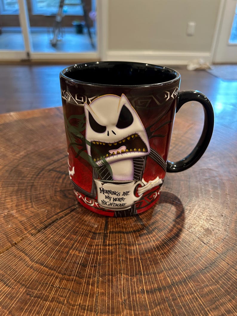 May include: Black ceramic mug featuring Jack Skellington from The Nightmare Before Christmas. The mug has a black handle and interior, with a design of Jack in a red and black background. Text on the mug reads "Mornings are my worst nightmare."