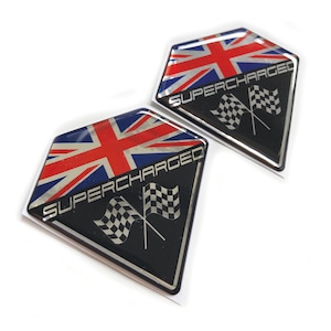 May include: Two diamond-shaped car emblems. The top half features the Union Jack flag design, with red, white, and blue colors. The bottom half is black with the word "SUPERCHARGED" and two checkered flags. The emblems are glossy.