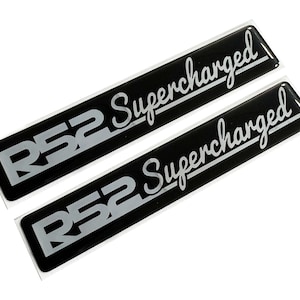 Puede incluir: Dos pegatinas rectangulares negras con el texto "R52 Supercharged" en blanco. Las pegatinas tienen un acabado brillante y se muestran sobre un fondo blanco. El texto está en una fuente estilizada.