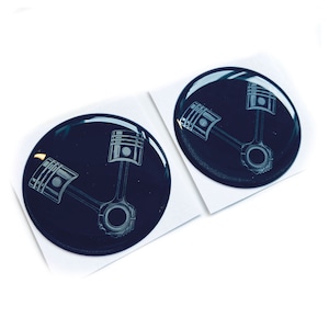 May include: Two glossy, dark blue, circular stickers. Each sticker features a detailed, light gray illustration of a piston and connecting rod. The stickers are on white backing paper, suitable for automotive enthusiasts or mechanics.