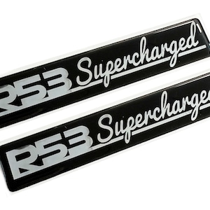 May include: Two black and silver car emblems with the text "R53 Supercharged" in a cursive font.