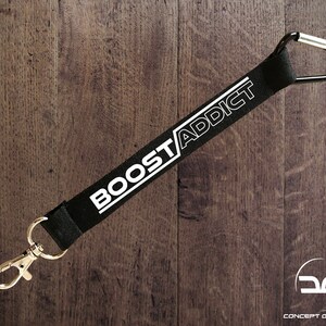 Puede incluir: Lanyard negro con texto blanco "BOOST/ADDICT" y un mosquetón plateado.
