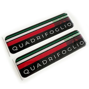 Puede incluir: Dos pegatinas en blanco y negro con un diseño de bandera italiana roja, blanca y verde. Las pegatinas dicen "Quadrifoglio" en letras blancas.