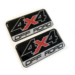 Puede incluir: Dos pegatinas negras y plateadas 4x4 Off Road con una 'X' roja en el centro. Las pegatinas tienen un acabado brillante y son rectangulares. El texto es en negrita y sin serifa.