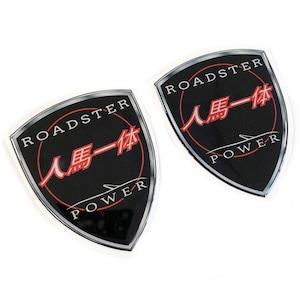 Pode incluir: Dois adesivos pretos e prateados com o emblema Roadster Power. Cada adesivo tem a forma de um escudo com a palavra "ROADSTER" no topo, caracteres japoneses no centro e "POWER" na parte inferior. Os adesivos estão em um fundo branco.