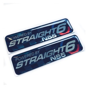 Könnte beinhalten: Zwei blaue und silberne 3D-Aufkleber mit dem Text "Powered by Straight 6 N55" in weißer Schrift. Die Aufkleber haben ein kariertes Muster und einen roten und blauen Kreis um die Zahl 6.