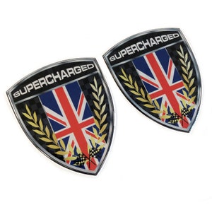 May include: Two silver and black shield-shaped stickers with the text "SUPERCHARGED" in white letters. The stickers feature a Union Jack flag with gold laurel wreaths and checkered racing flags.