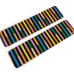Puede incluir: Dos pegatinas rectangulares con un patrón de rayas de colores. Las rayas son negras, naranjas, amarillas, verdes, rosas y azules.