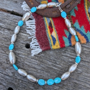 May include: A turquoise and silver necklace with oval-shaped beads. The necklace is lying on a wooden surface with a colorful rug and a horseshoe in the background.