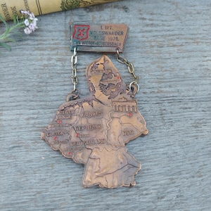 May include: A bronze-colored map of Germany with red dots marking the locations of cities. The map is attached to a chain with a small metal tag that reads "1. Int. Volkswander Tage 1970.  SVG Nieder-Liebersbach."