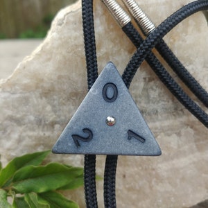 May include: A black bolo tie with a gray triangular pendant featuring the numbers 2017. The pendant has a silver stud in the center.