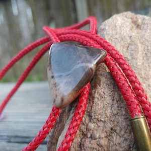 May include: A red braided bolo tie with a brown and grey stone pendant. The stone is triangular in shape and has a natural, polished finish.
