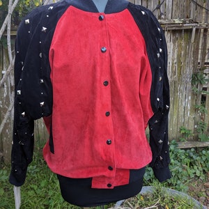May include: A red suede bomber jacket with black suede sleeves and silver pyramid studs. The jacket has a black ribbed collar and cuffs.