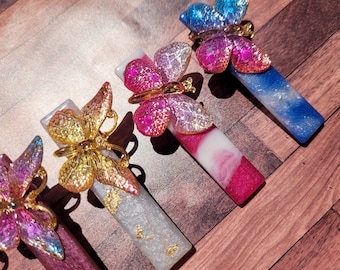 Puede incluir: Conjunto de tapones decorativos para botellas con diseños de mariposas. Cada tapón presenta una base rectangular con un acabado brillante y colorido, coronada por una mariposa detallada en dorado, rosa y azul.
