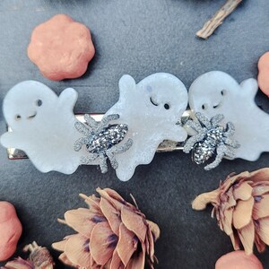 May include: Three white ghost-shaped hair clips with silver glitter spiders. The ghosts have black eyes and a black smile.