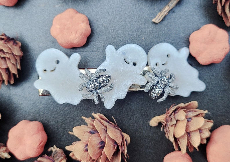 May include: Three white ghost-shaped hair clips with silver glitter spiders. The ghosts have black eyes and a black smile.