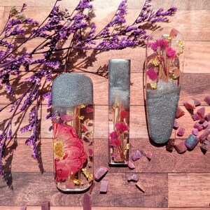May include: Three silver hair clips with pressed flowers and gold leaf accents. The clips are on a wooden surface with purple flowers and pink and purple glitter.