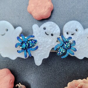 May include: Three white ghost-shaped hair clips with blue glitter spiders. The ghosts have black eyes and a black smile.