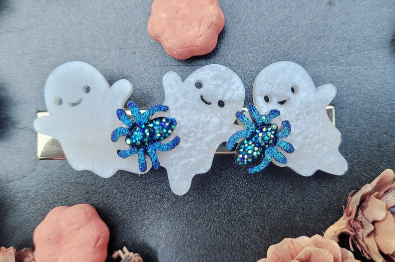 May include: Three white ghost-shaped hair clips with blue glitter spiders. The ghosts have black eyes and a black smile.