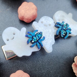 May include: Three white ghost-shaped hair clips with iridescent blue and green spiders attached to each one.