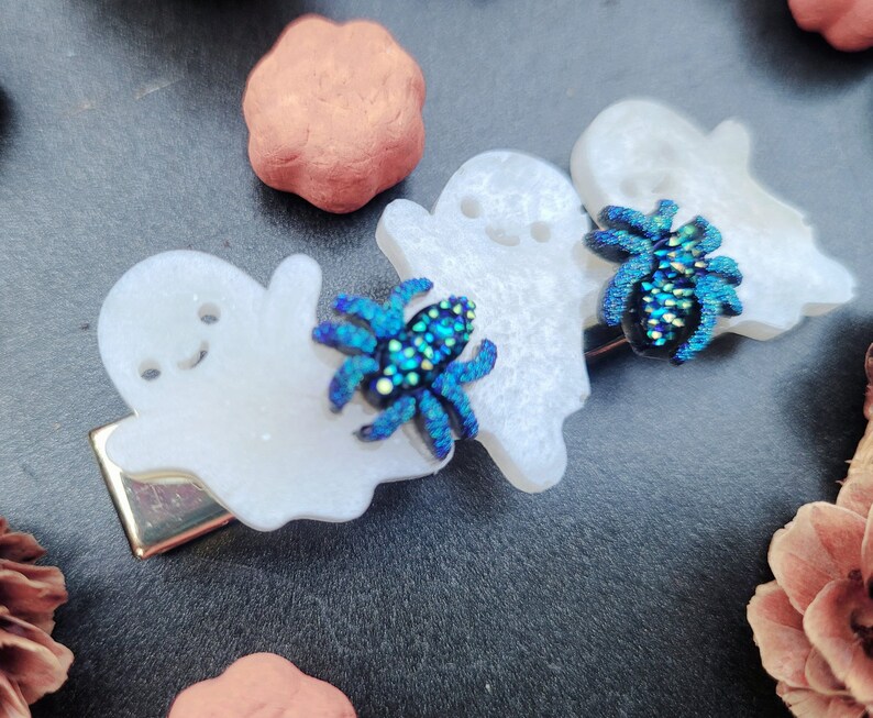 May include: Three white ghost-shaped hair clips with iridescent blue and green spiders attached to each one.
