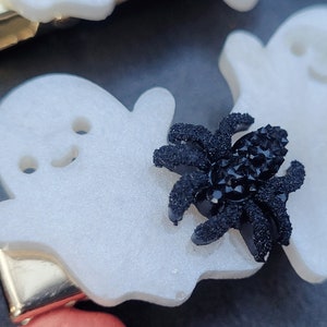 May include: A white ghost-shaped hair clip with a black sparkly spider on top. The clip is made of plastic and has a gold metal bar on the back.