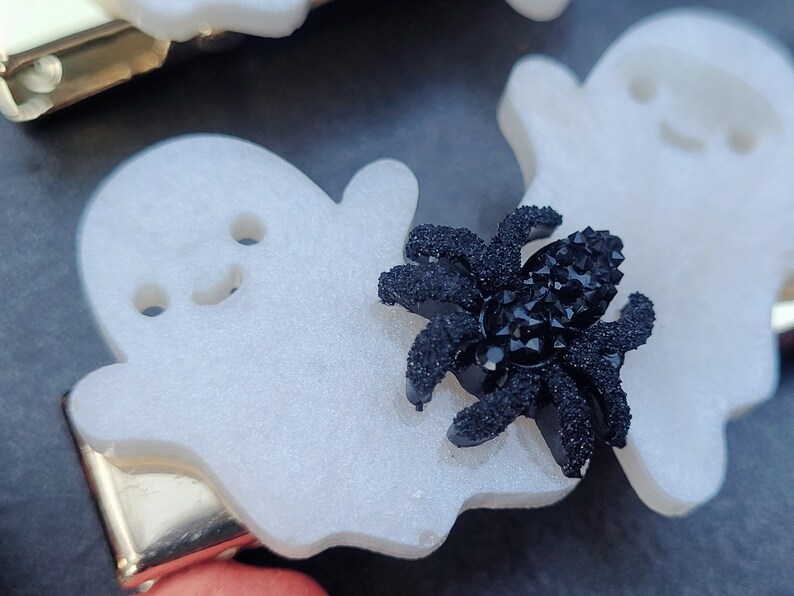 May include: A white ghost-shaped hair clip with a black sparkly spider on top. The clip is made of plastic and has a gold metal bar on the back.