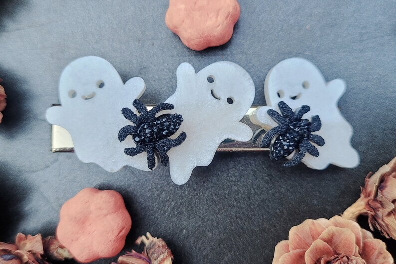 May include: Three white ghost-shaped hair clips with black glitter spiders. The ghosts have a smiling face and are translucent.