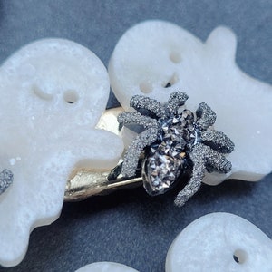 May include: A white ghost-shaped hair clip with a sparkly black spider embellishment. The clip is made of plastic and has a silver metal bar.