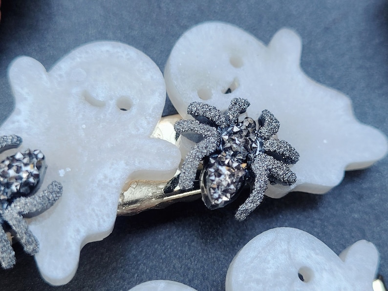 May include: A white ghost-shaped hair clip with a sparkly black spider embellishment. The clip is made of plastic and has a silver metal bar.