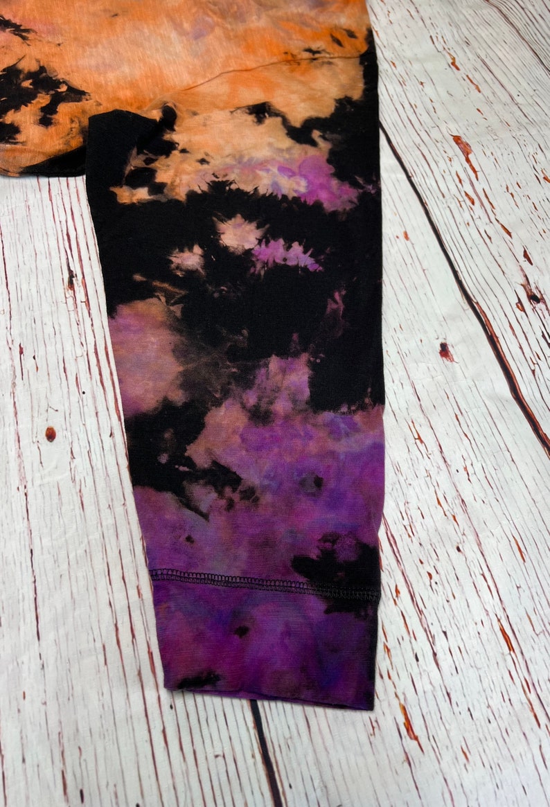 Op de afbeelding: Een zwart longsleeve shirt met een tie-dye patroon in tinten oranje, paars en zwart. Het shirt heeft een geribbelde manchet en is gemaakt van een zachte, comfortabele stof.