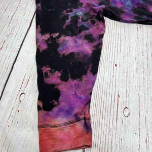 Op de afbeelding: Een shirt met lange mouwen met een tie-dye patroon in de kleuren zwart, paars en roze. Het shirt heeft een rode en roze zoom.