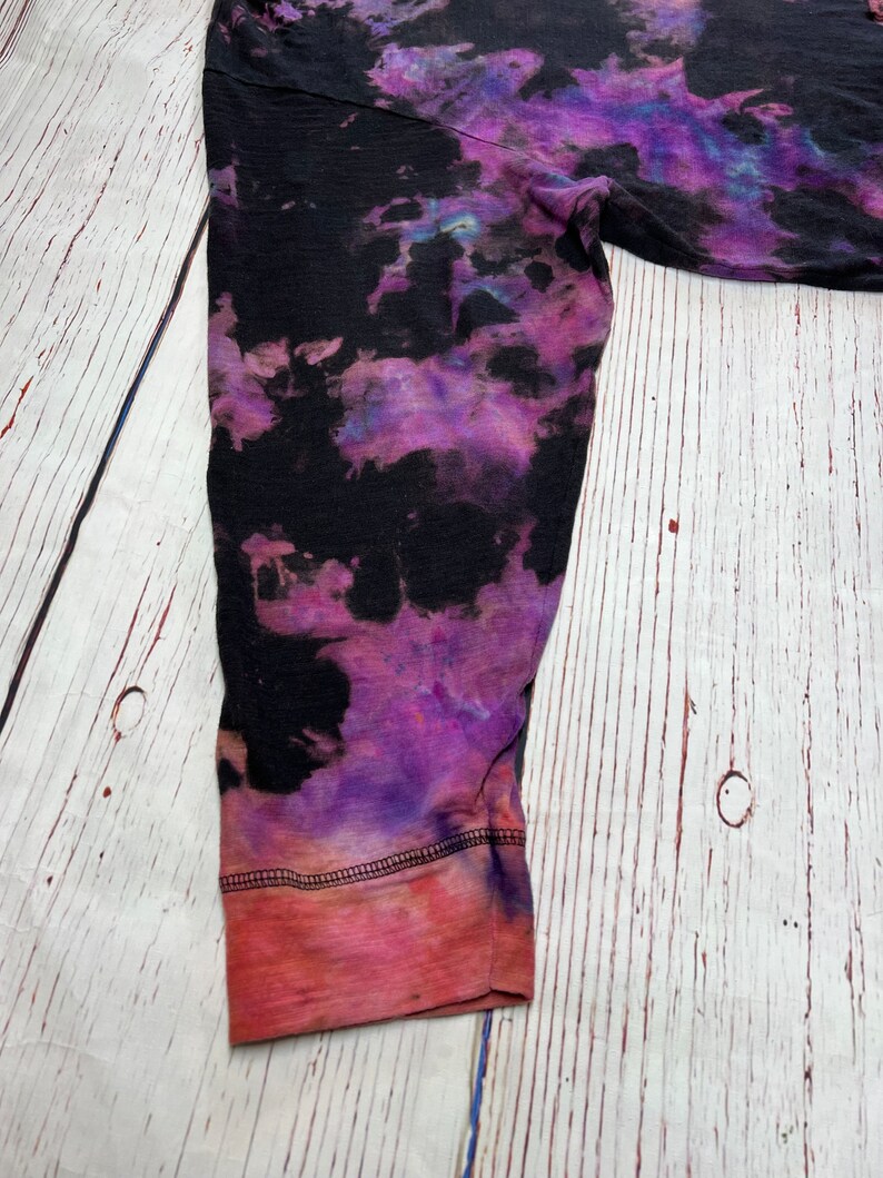 Op de afbeelding: Een shirt met lange mouwen met een tie-dye patroon in de kleuren zwart, paars en roze. Het shirt heeft een rode en roze zoom.
