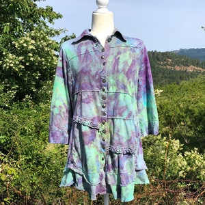 May include: A tie-dye button-up shirt with a ruffled hem. The shirt is a mix of purple, green, and blue colors. The shirt is on a white mannequin.
