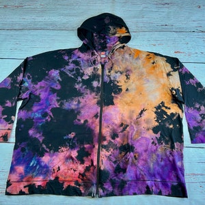 Op de afbeelding: Een zwarte hoodie met rits met een tie-dye patroon in tinten paars, oranje en roze.
