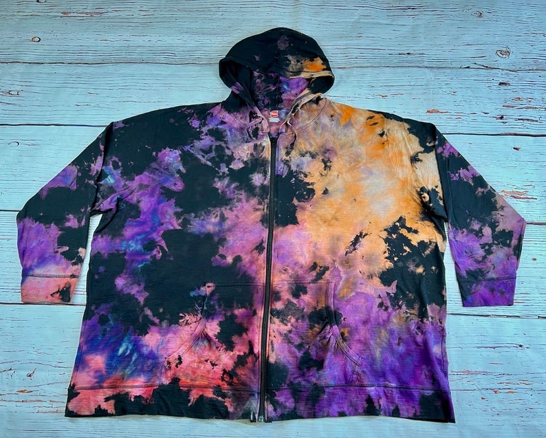 Op de afbeelding: Een zwarte hoodie met rits met een tie-dye patroon in tinten paars, oranje en roze.