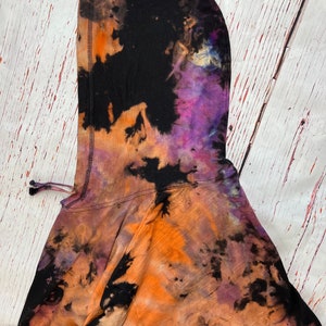 Op de afbeelding: Een zwart hoodie met een tie-dye patroon in tinten oranje, paars en zwart.