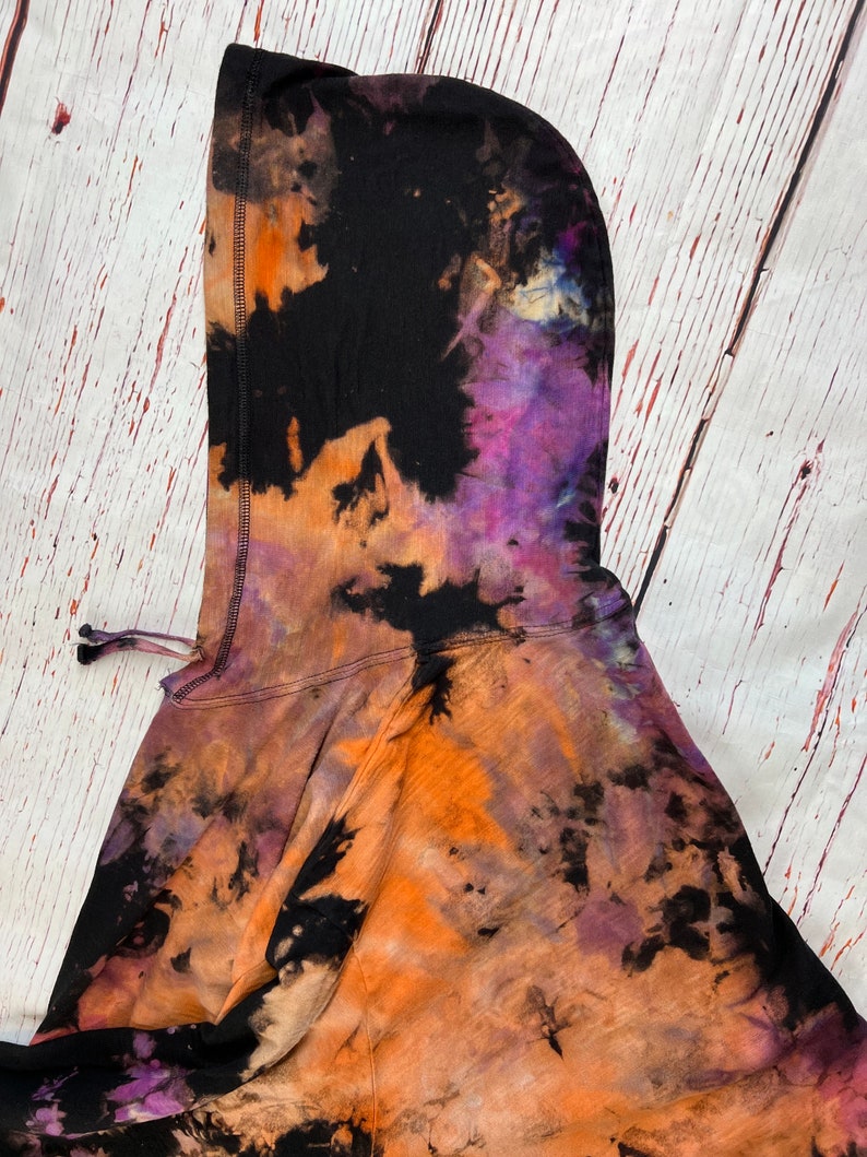 Op de afbeelding: Een zwart hoodie met een tie-dye patroon in tinten oranje, paars en zwart.