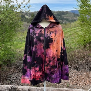 Op de afbeelding: Een zwarte hoodie met rits en een tie-dye patroon in tinten paars, oranje en zwart. De hoodie staat op een wit mannequin en heeft een capuchon.